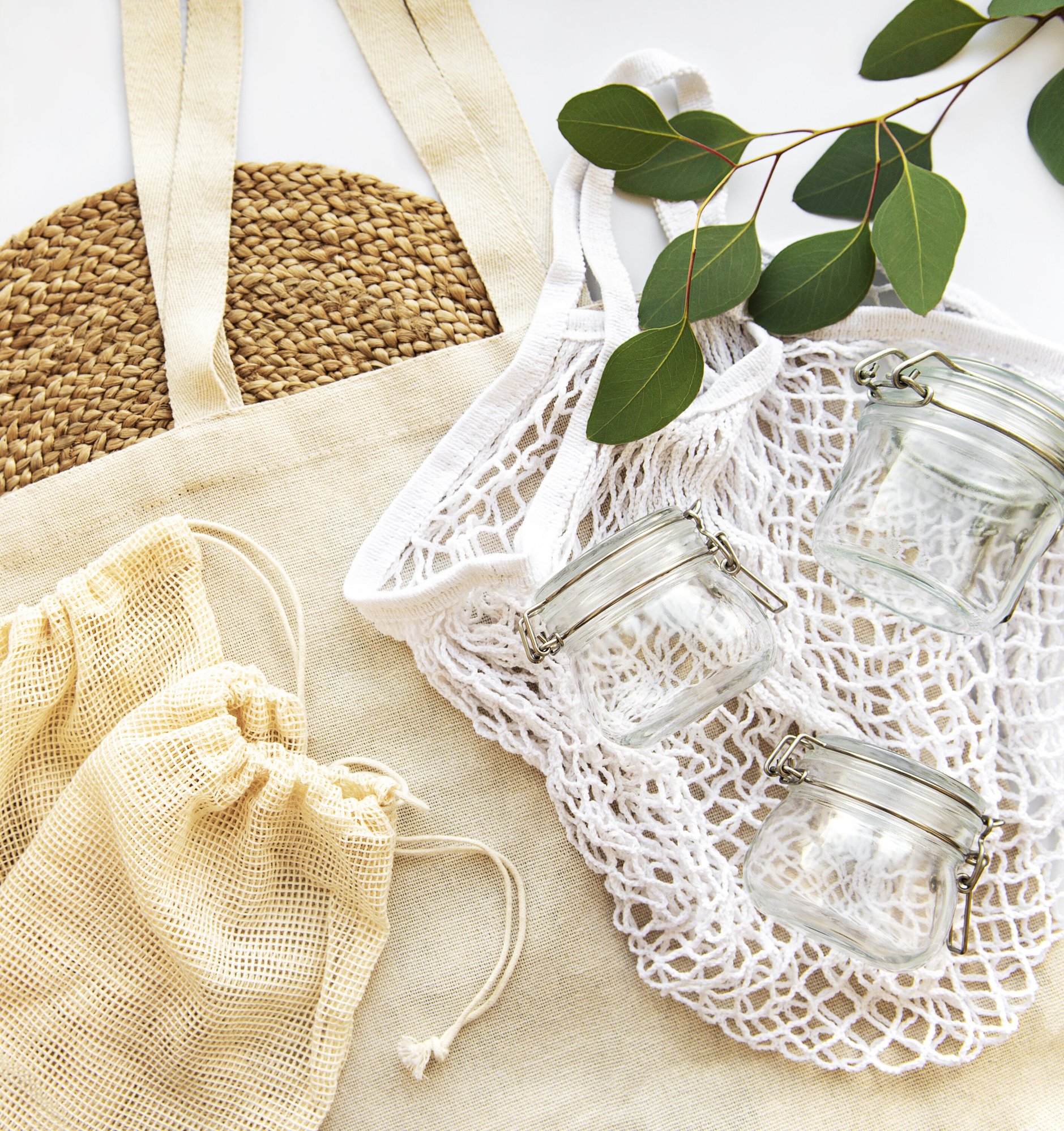 Flat lay of reusable fabric bags, small mesh pouches, and clear glass jars arranged with eucalyptus leaves: soft neutral textures and sustainable materials.