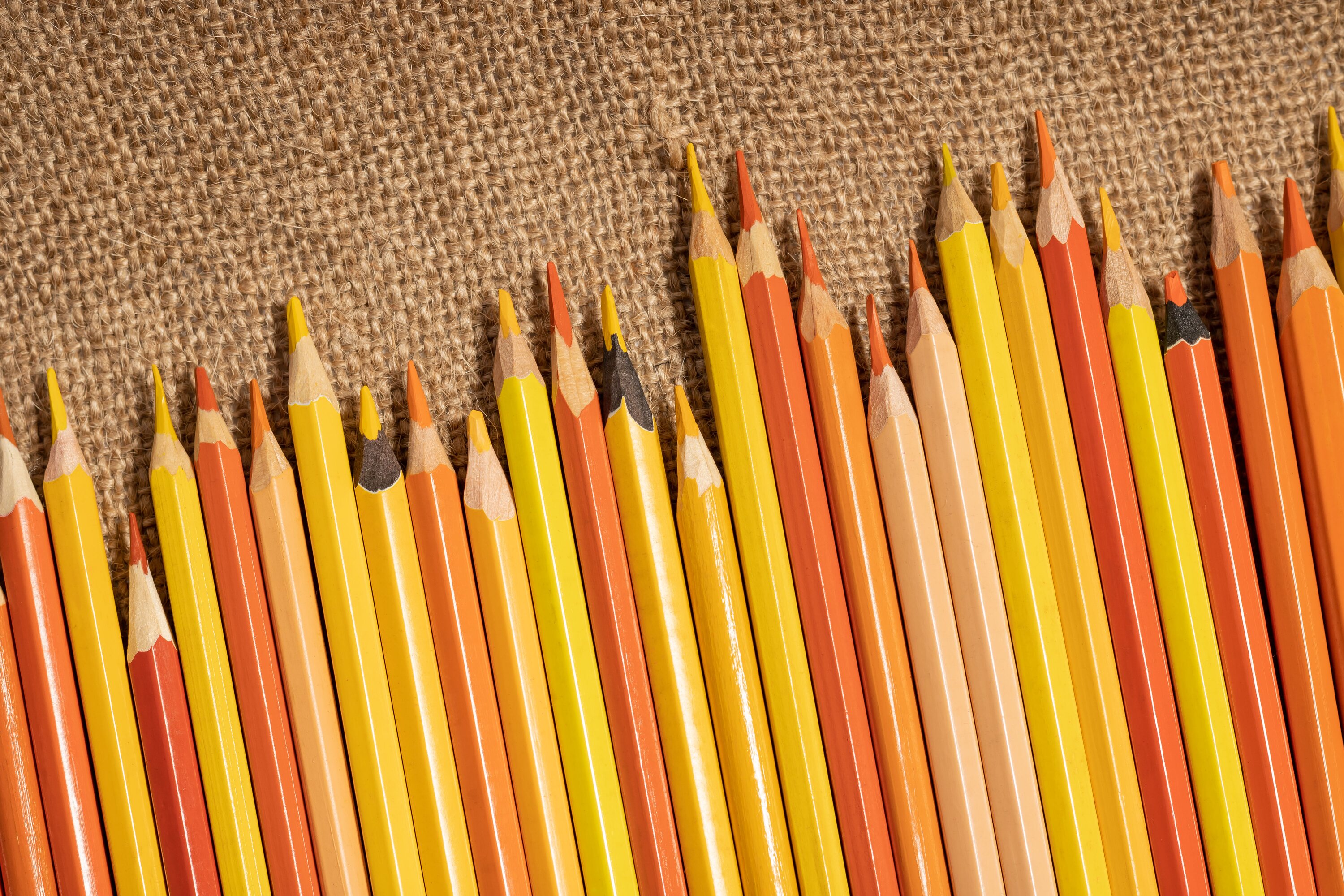 Row of orange and yellow colored pencils scattered across burlap fabric — warm tones and craft materials.